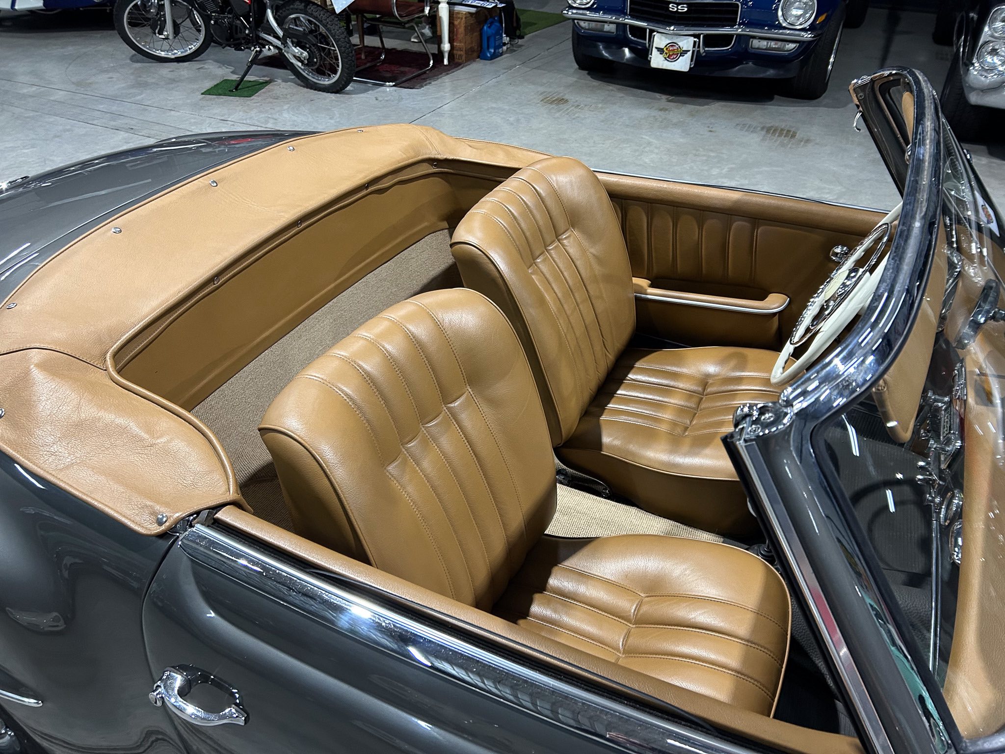 Open-top tan leather interior of a vintage convertible, two front seats and chrome details in a showroom setting with other classic cars in the background