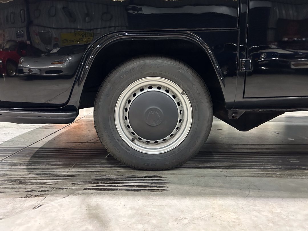 Side view of a black van’s wheel: silver steel rim with hubcap and tire, parked indoors on a concrete floor.