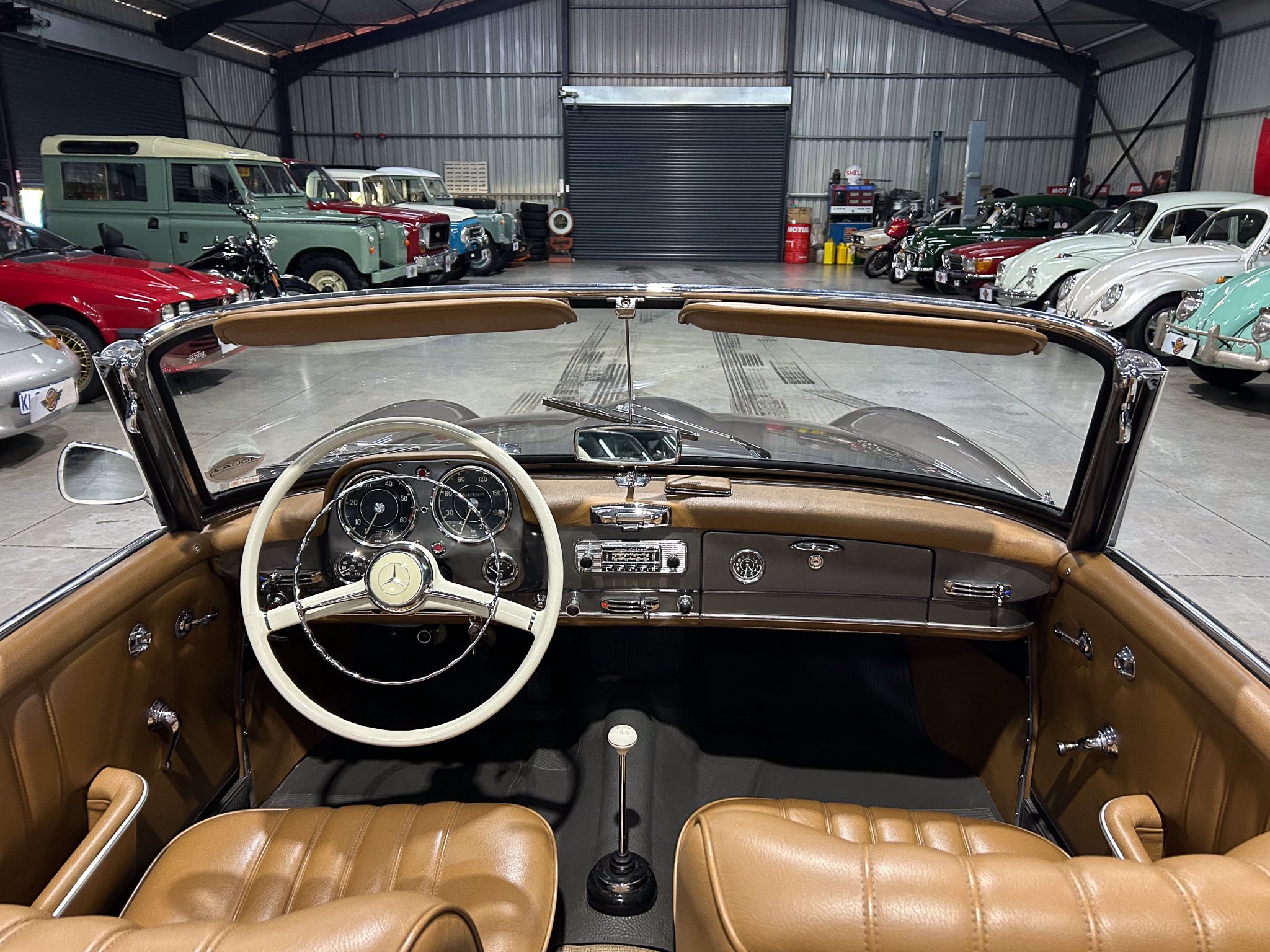 From the driver's seat of a tan leather vintage convertible, dashboard gauges visible as a showroom full of classic cars lines the sides behind.