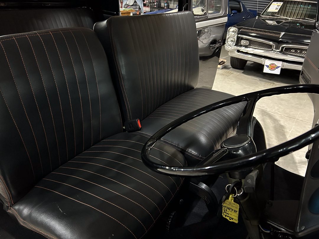 Front bench seat and steering wheel of a vintage black leather car with orange stitching, shown in a showroom with other classic cars in the background.