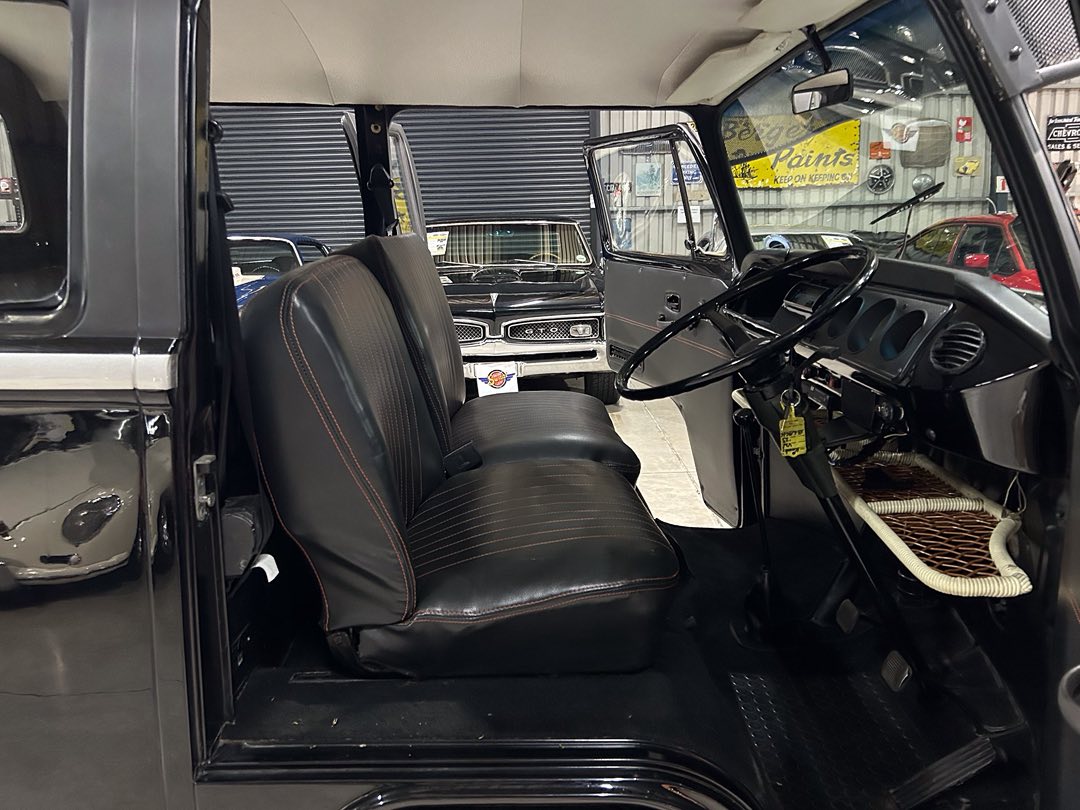 Interior of a vintage pickup truck with black vinyl bench seats, red-stitched seams, and a large classic steering wheel, viewed from the open side door.