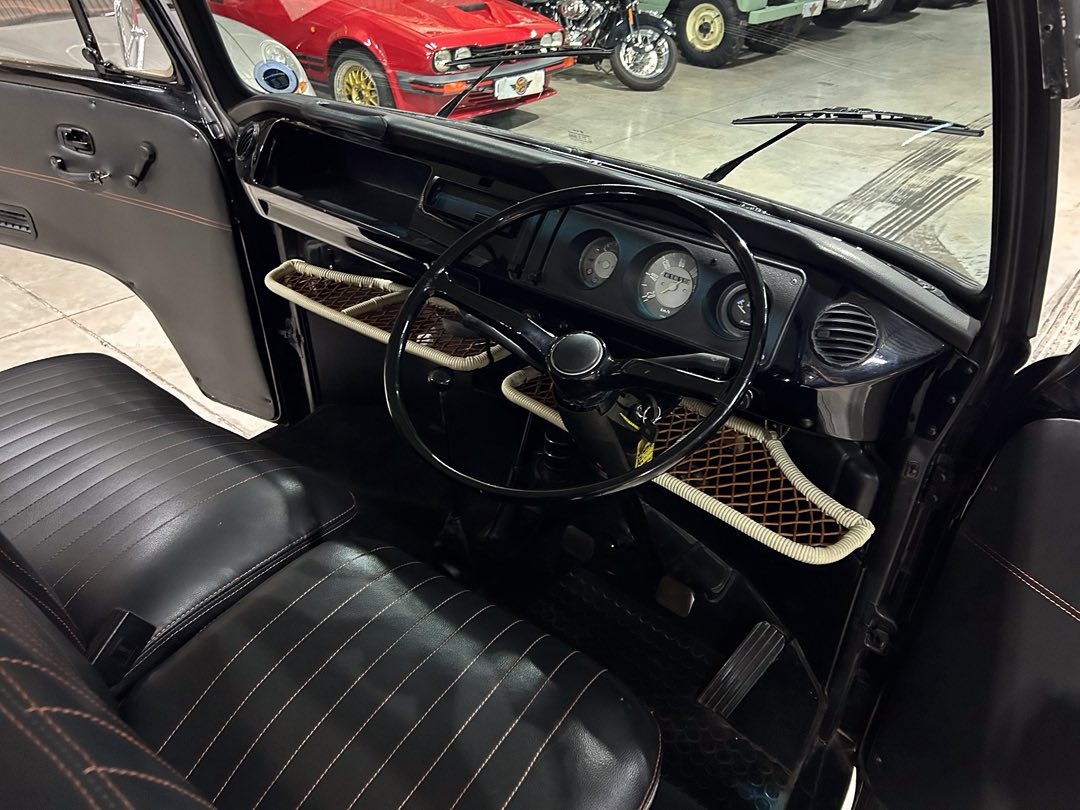 Interior of a vintage black car with leather seats, a large circular steering wheel, and analog dash gauges visible from the driver’s side edge. The dash has a woven storage net below. Other classic cars are seen in the background in a showroom.