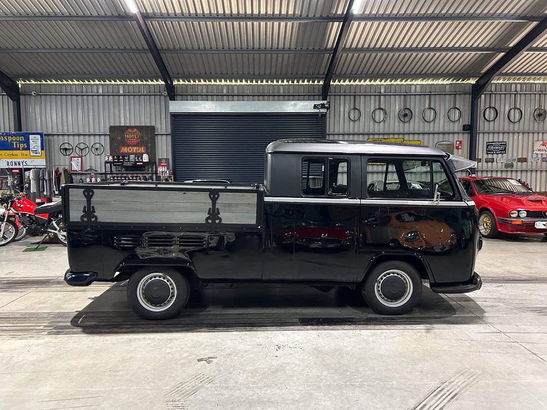 Classic black vintage panel van with a wooden flatbed, parked inside a showroom with other vehicles in the background.