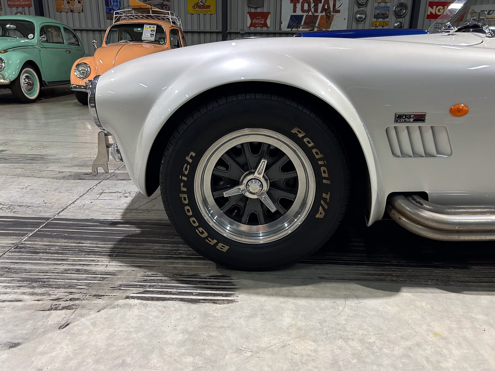 Close-up of a silver classic car's front wheel and fender, with a black tire and chrome hubcap, in a showroom with pastel cars in the background.