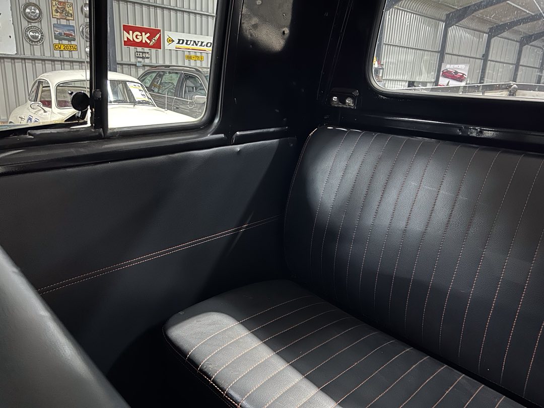 Interior of a vintage car with black leather seats accented by light brown stitching, view of a garage with classic cars outside.