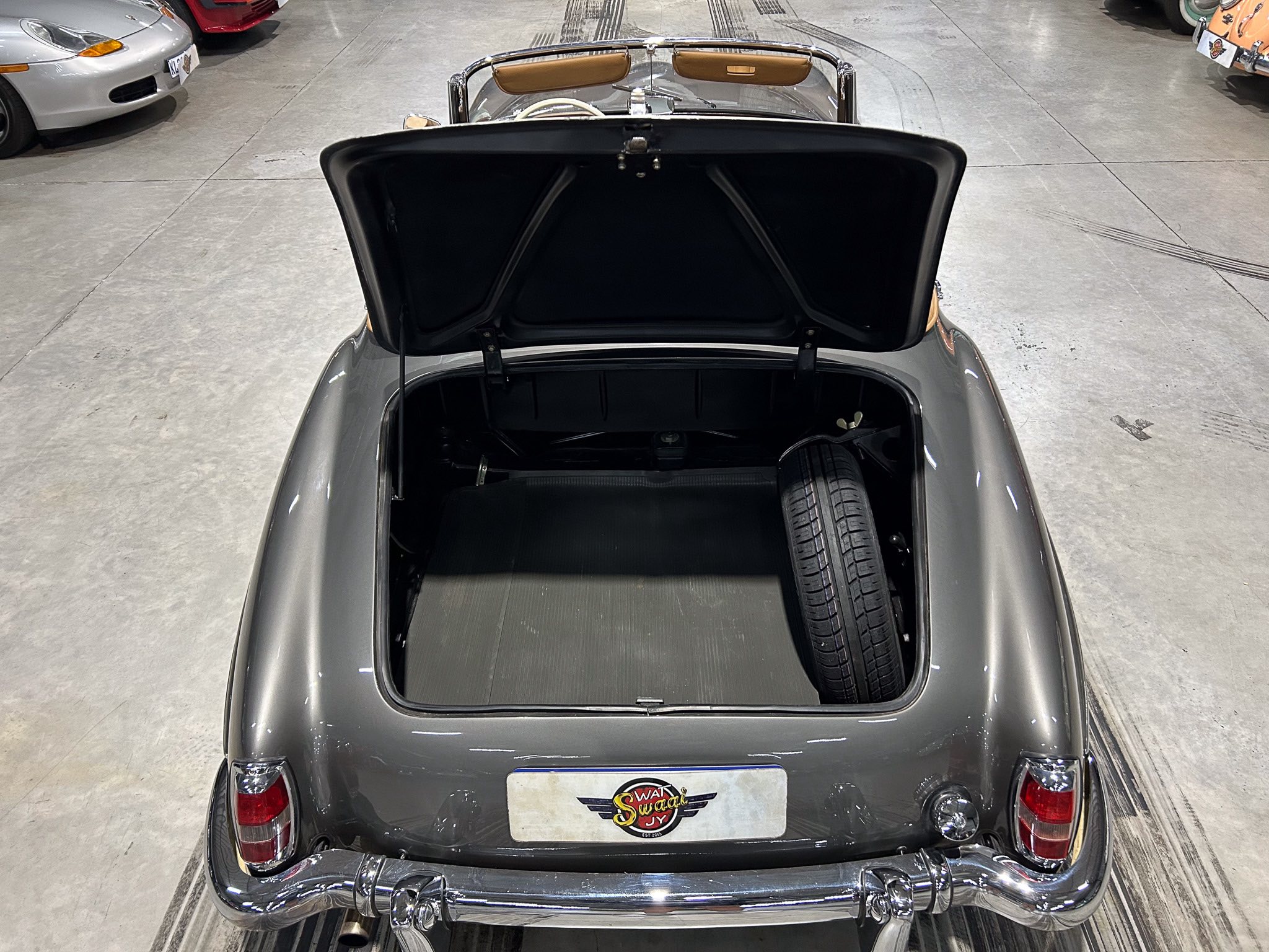Open trunk of a gray vintage car in a showroom, with a spare tire visible in the trunk and chrome bumper details nearby