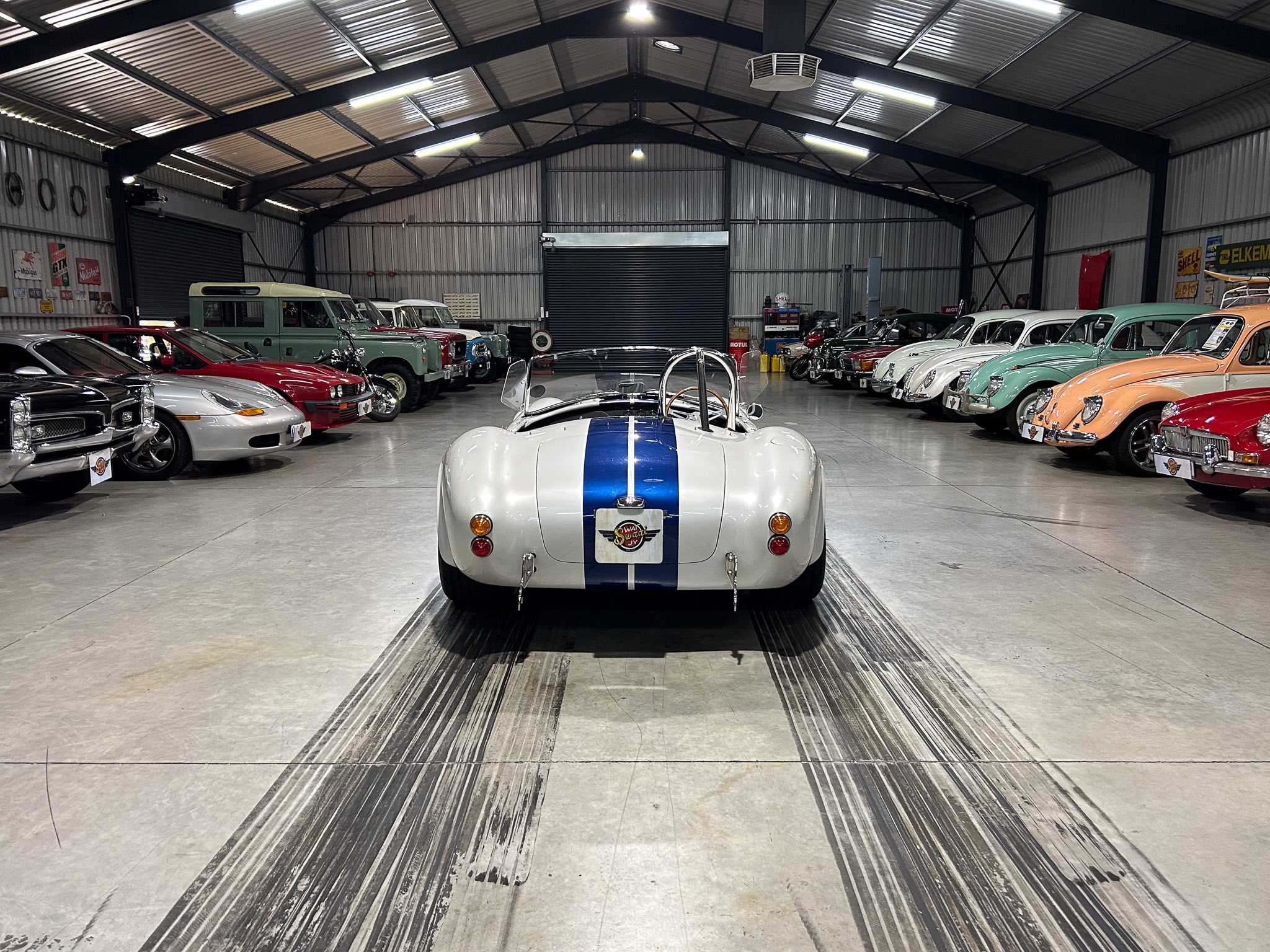 Classic car showroom with a white race car in the foreground and rows of vintage cars along both sides inside a metal warehouse.