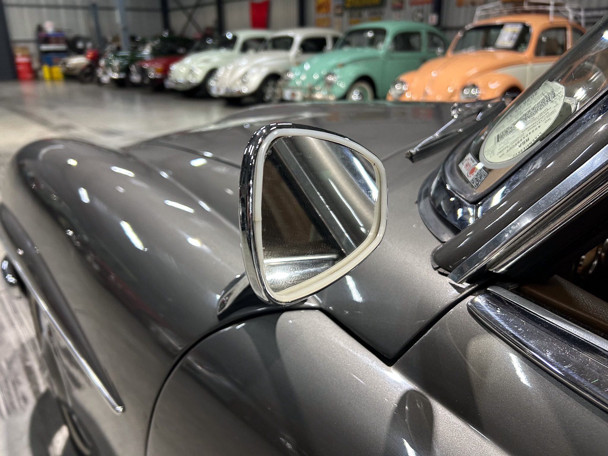 Close-up of a chrome side mirror on a dark car, with a row of vintage cars blurred in a showroom background.
