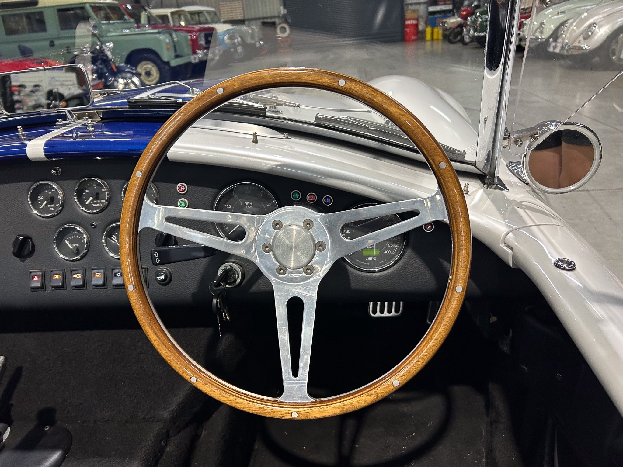 Inside a vintage car cockpit with a wood-rimmed steering wheel, chrome dashboard gauges, and a showroom of classic cars in the background.