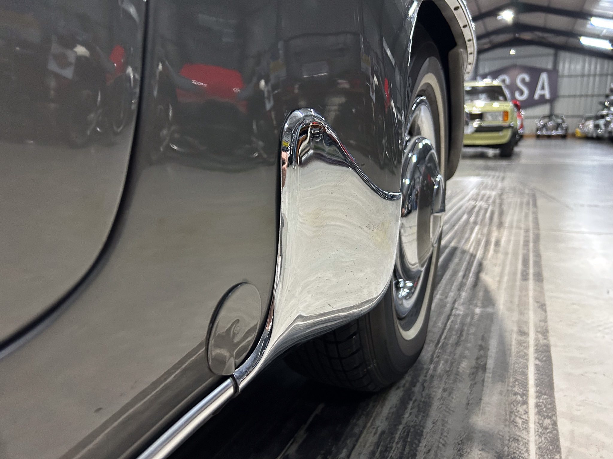 Close-up of a dark car's chrome side panel reflecting the showroom and other vehicles in the background.