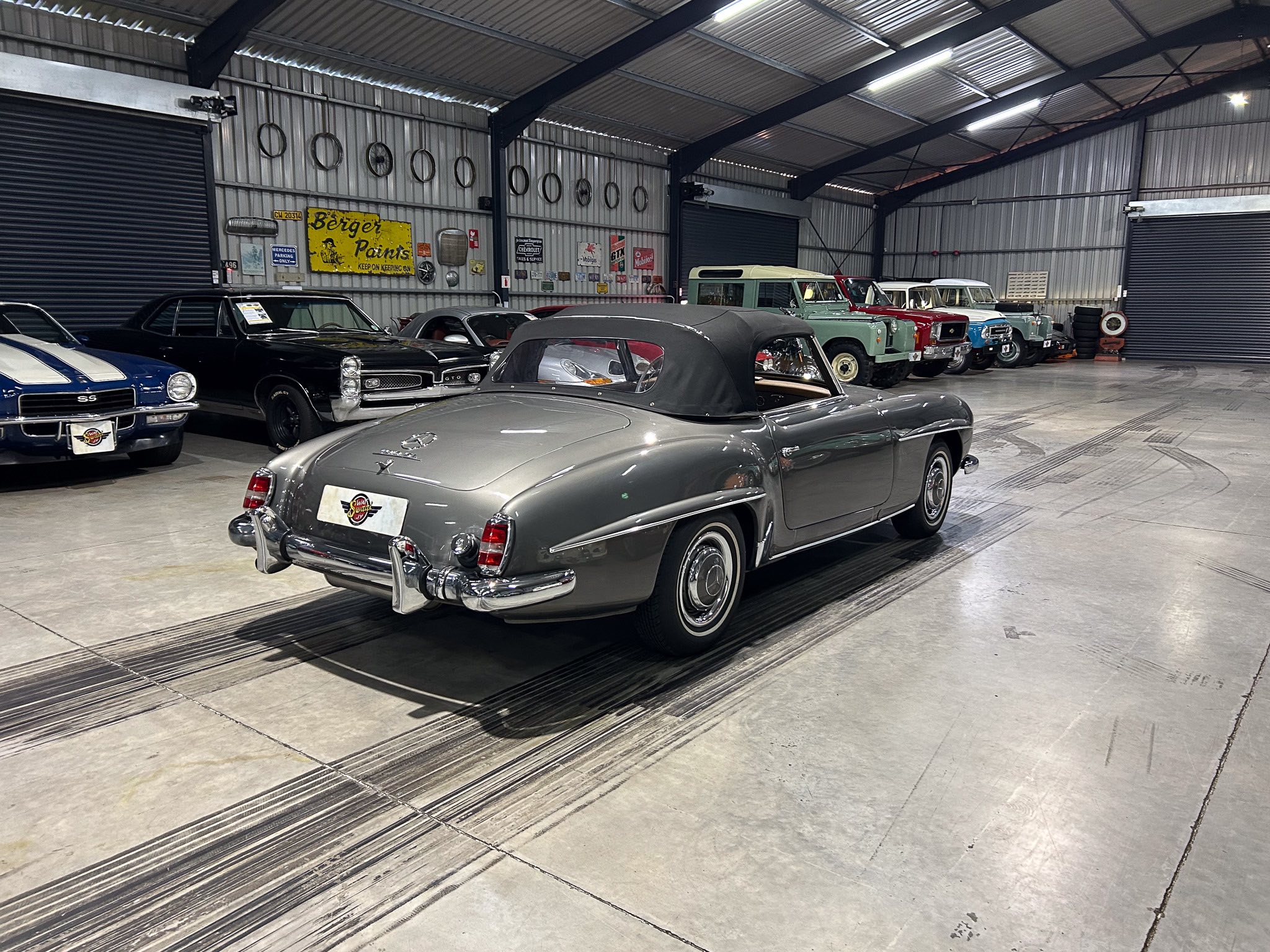 A silver vintage convertible in the foreground inside a metal-walled showroom, with classic cars lined up to the right and retro signage on the wall