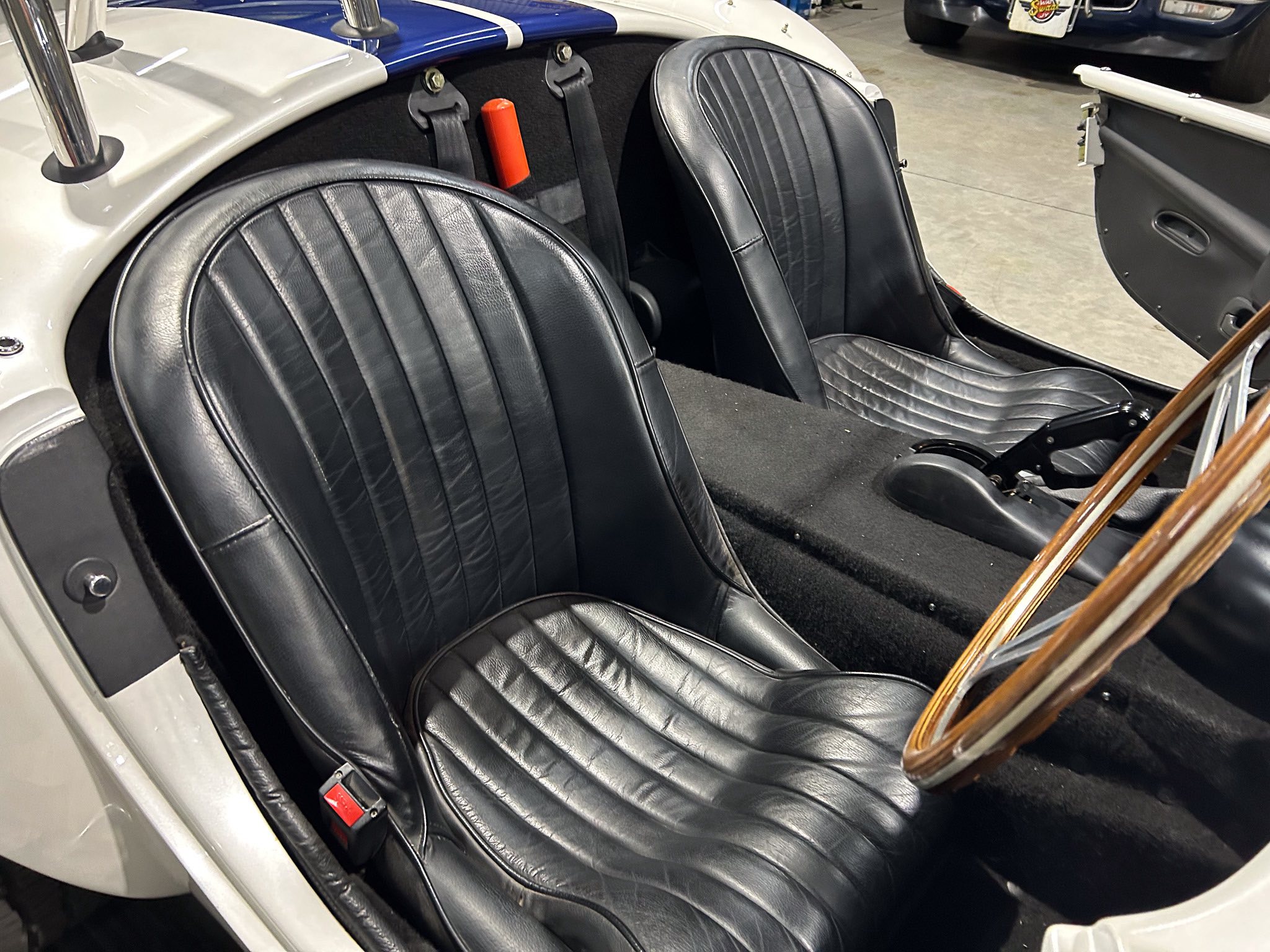 Two black leather seats in a vintage car interior with a red fire extinguisher mounted between them, and seat belts visible between the seats.