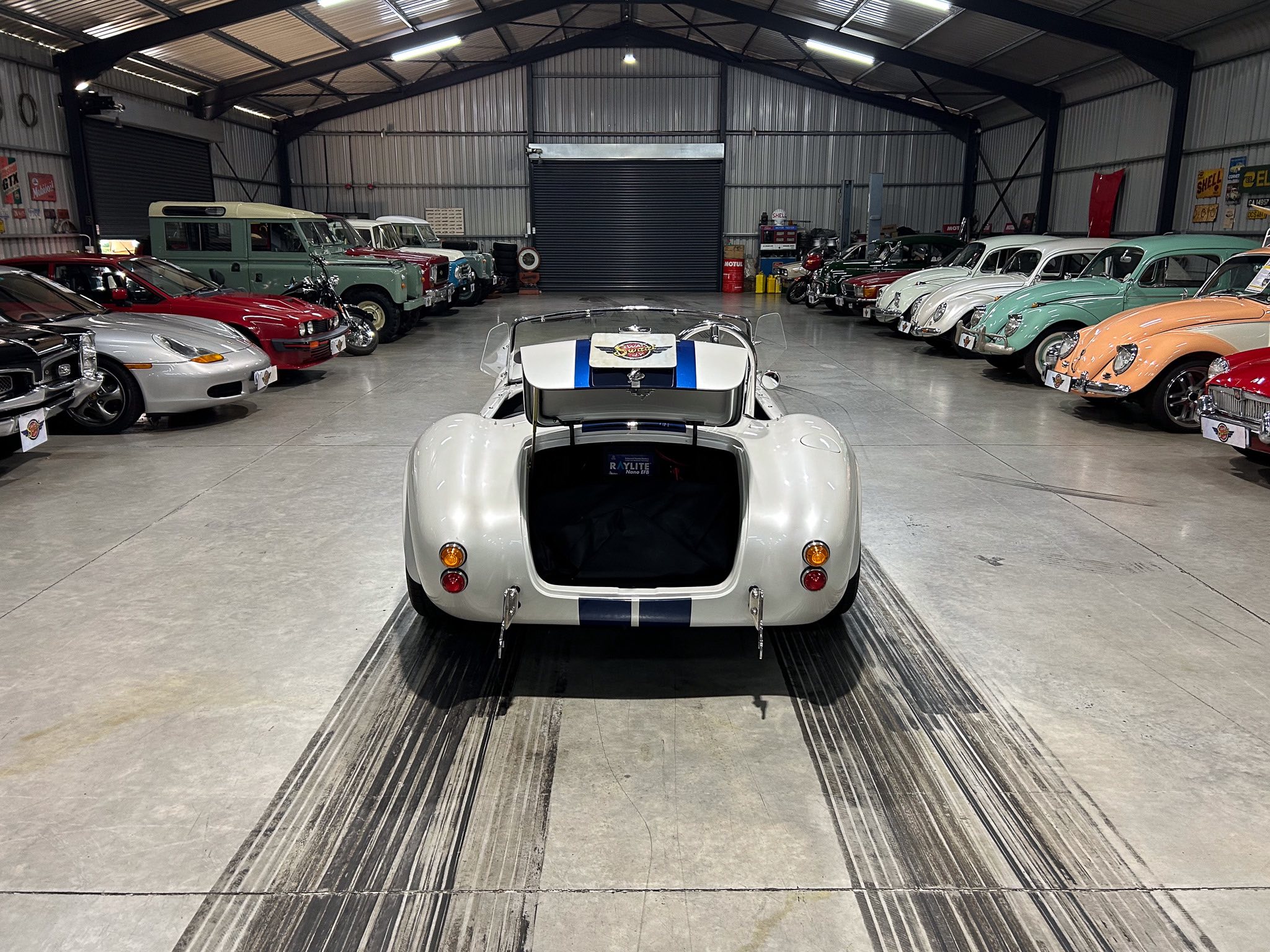 Interior of a large garage showroom filled with vintage and classic cars on both sides, viewed from the rear of a white race car with blue stripes.]