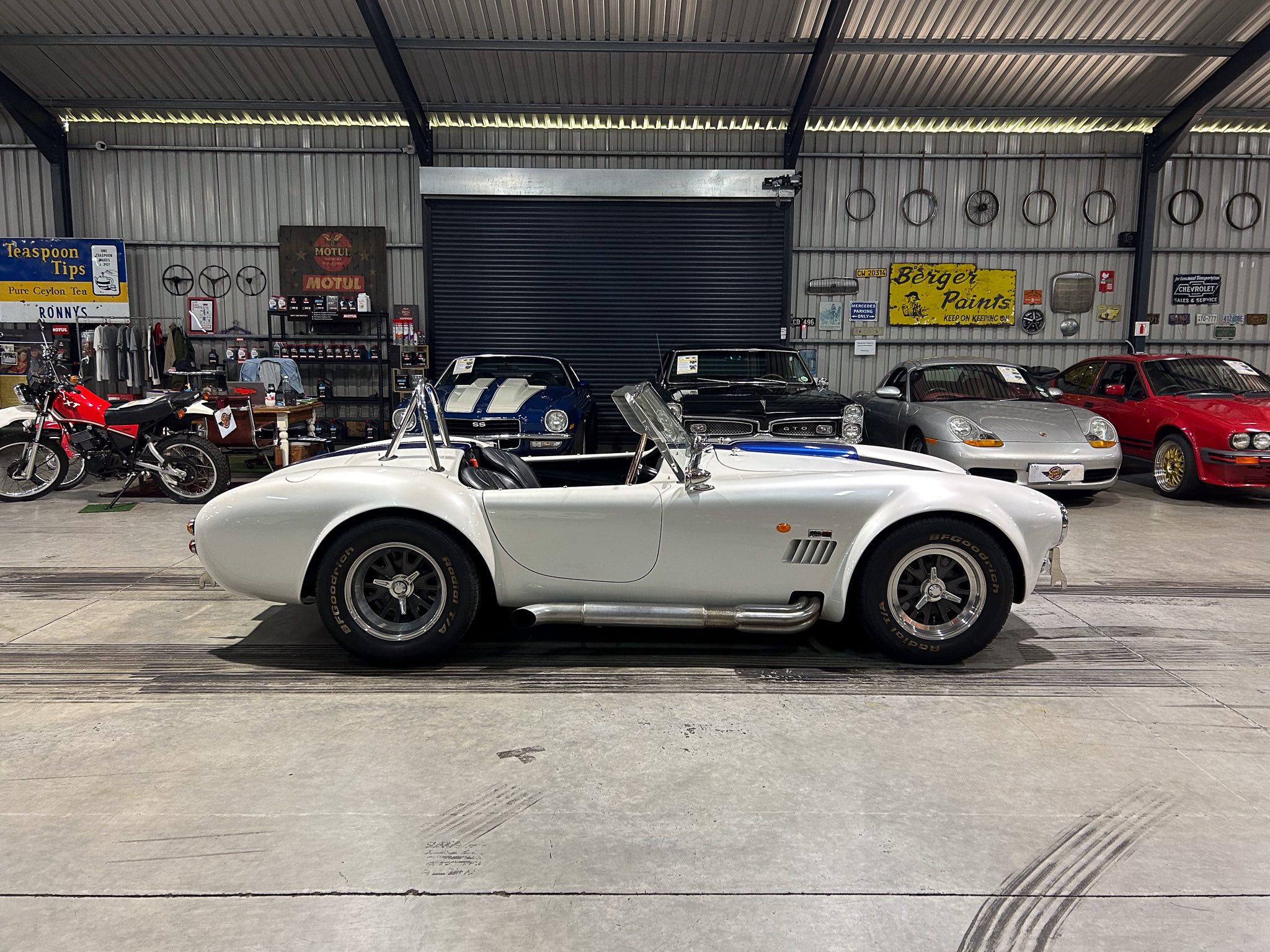 Indoor classic car showroom with a white convertible in the foreground and vintage signs on a metal-wall backdrop