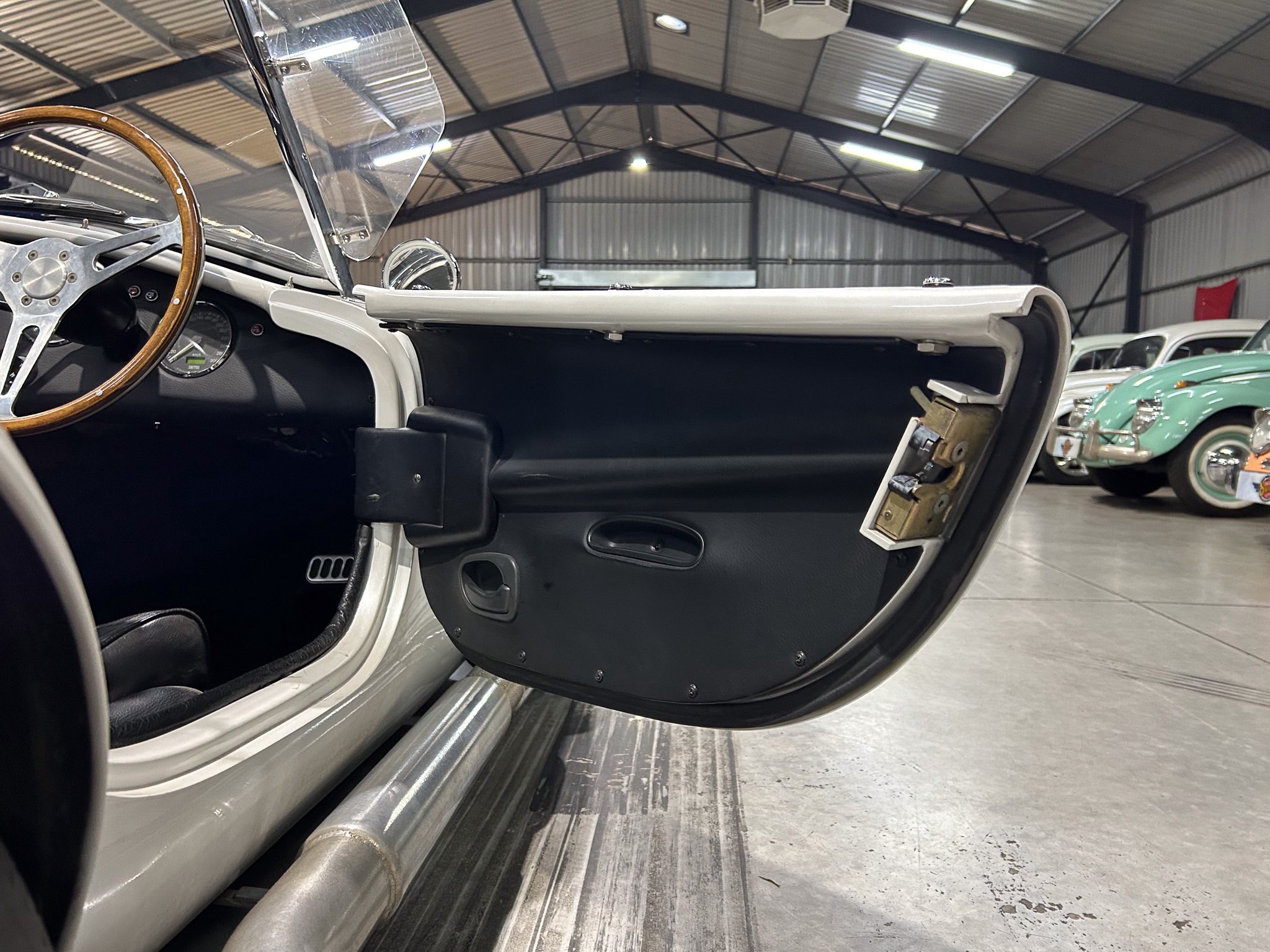 Open vintage convertible door interior: wood steering wheel, black dashboard, and chrome details in a showroom with other classic cars in background.