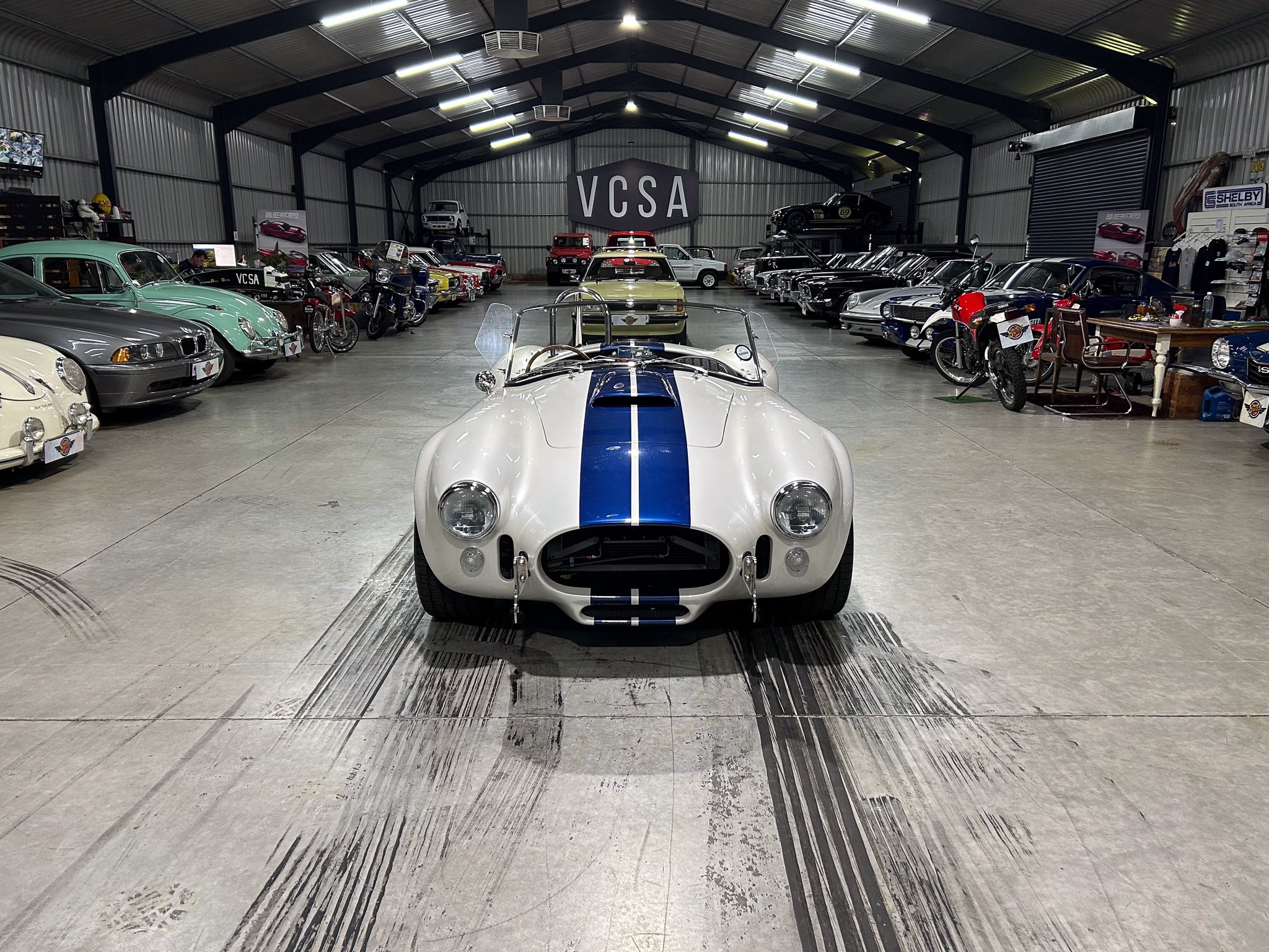 Classic car showroom or museum filled with vintage cars, seen from the front with a white racecar and blue stripe in the foreground