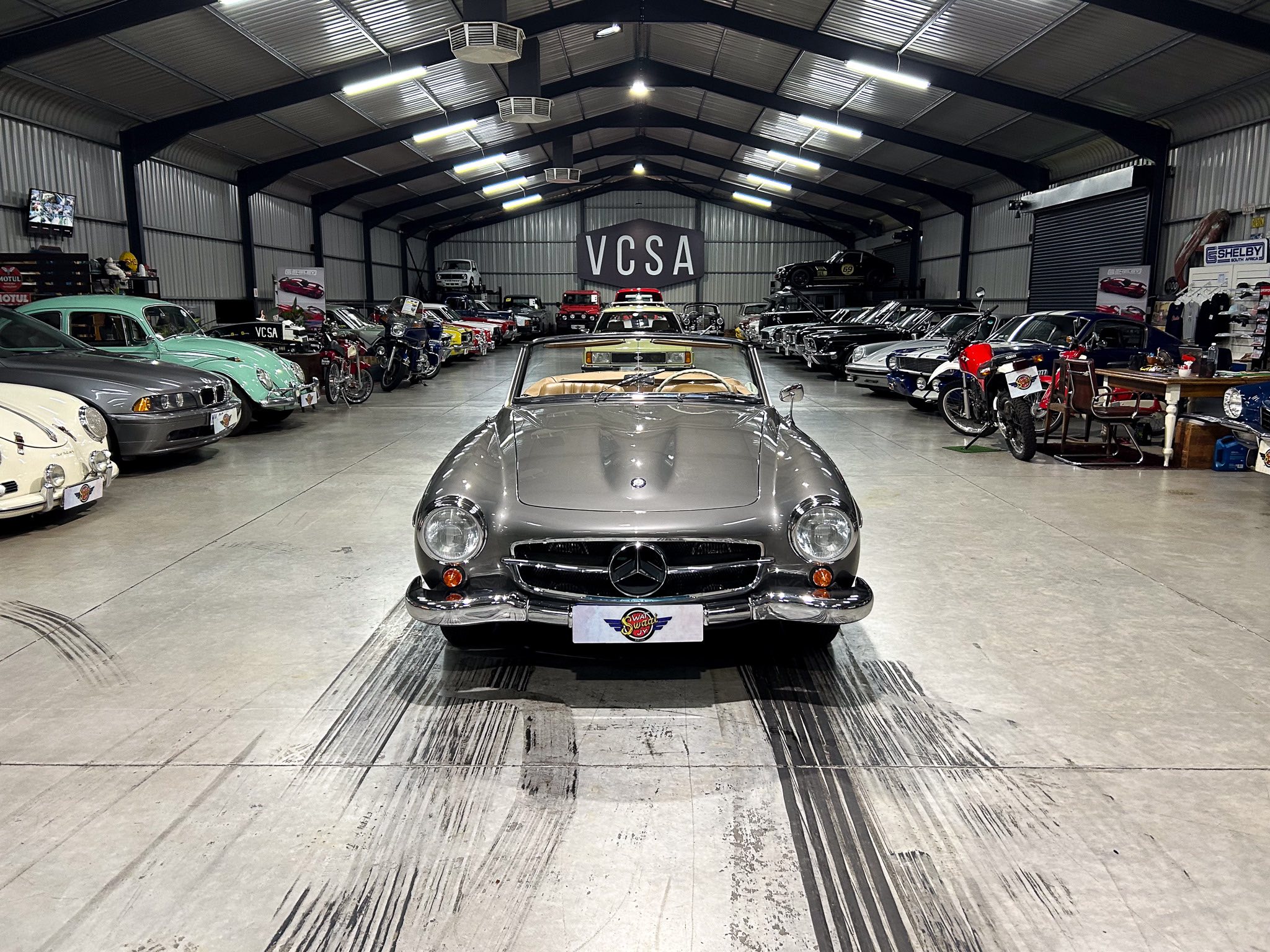 Front-and-center gray vintage Mercedes convertible inside a large indoor showroom filled with classic cars on both sides.