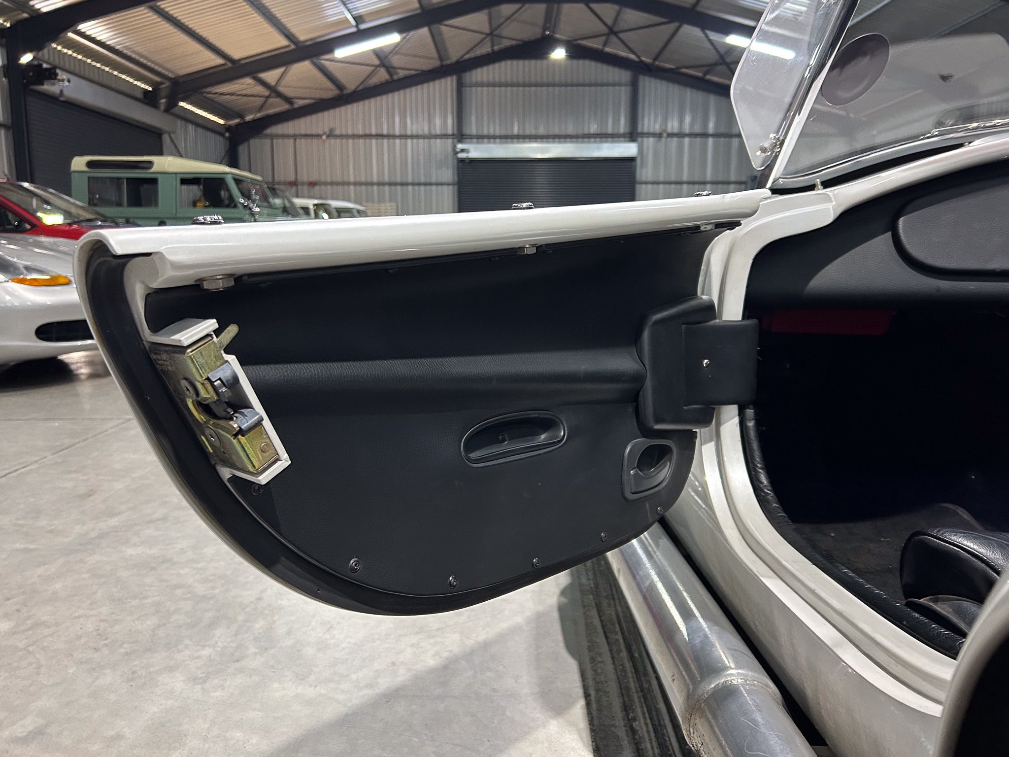 Open car door of a vintage convertible, exposing the black interior panel and chrome trim in a large garage with other vehicles in the background.
