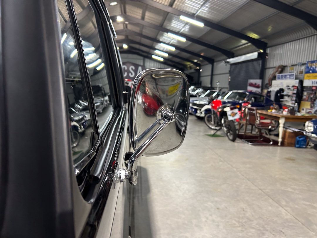Close-up of a chrome side mirror on a vintage car inside a showroom filled with classic vehicles and motorcycles in the background.