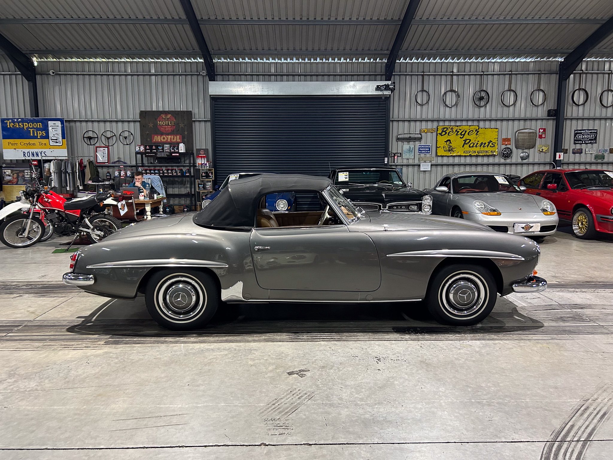Classic car showroom with a silver-gray convertible in the foreground and vintage signage on the back wall.