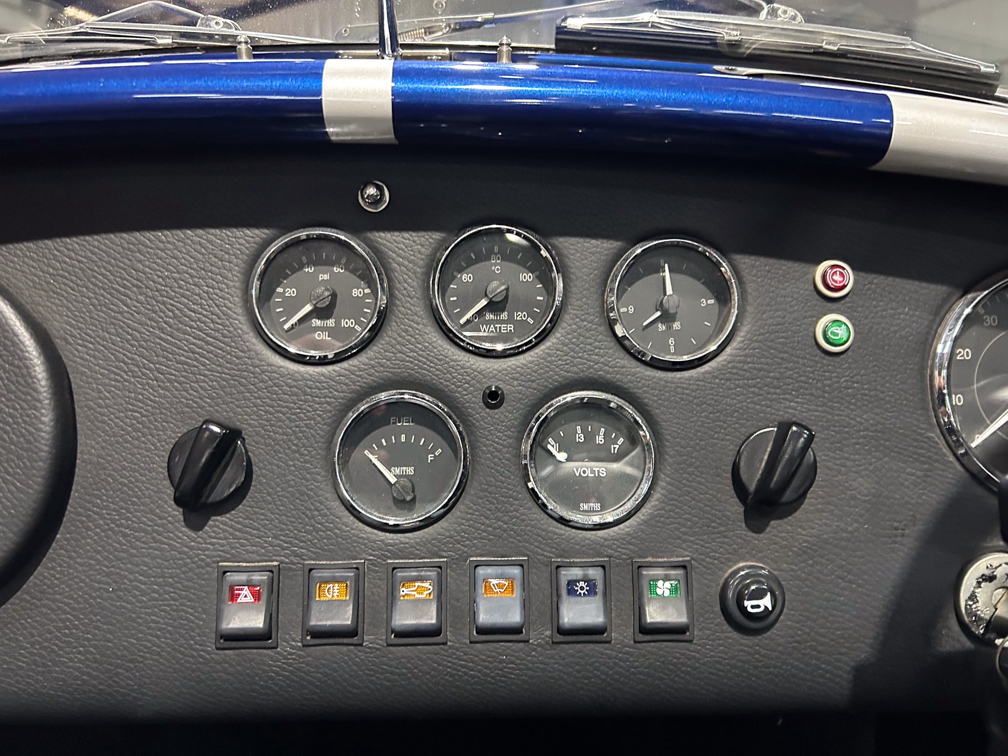 Close-up of a classic car instrument panel showing oil, water, and volt gauges with a row of indicator switches beneath.