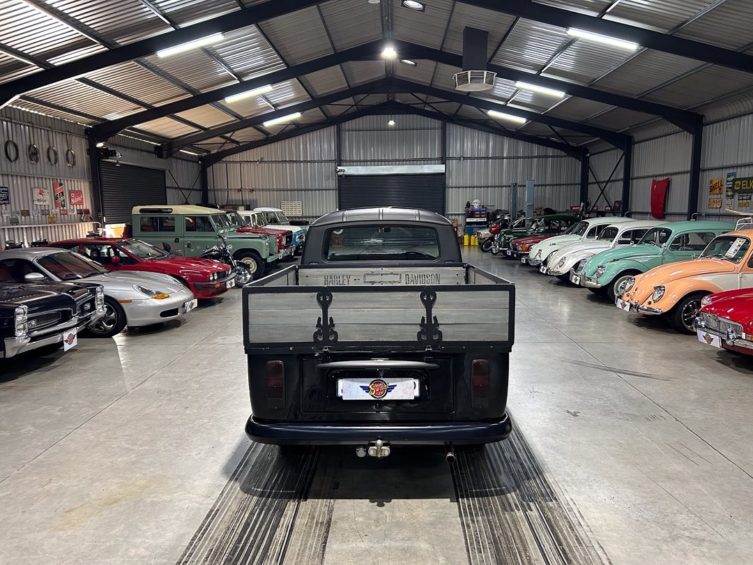 Rear view of a black vintage pickup with a wooden tailgate in a large showroom filled with colorful classic cars on both sides.