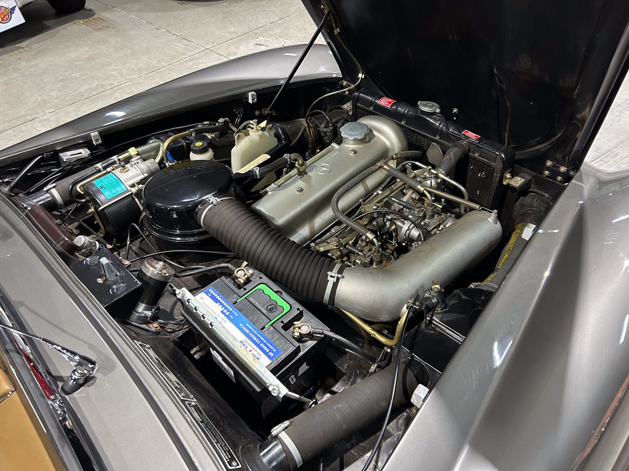 Open car hood revealing a classic Mercedes engine bay with a silver engine block and numerous hoses and components