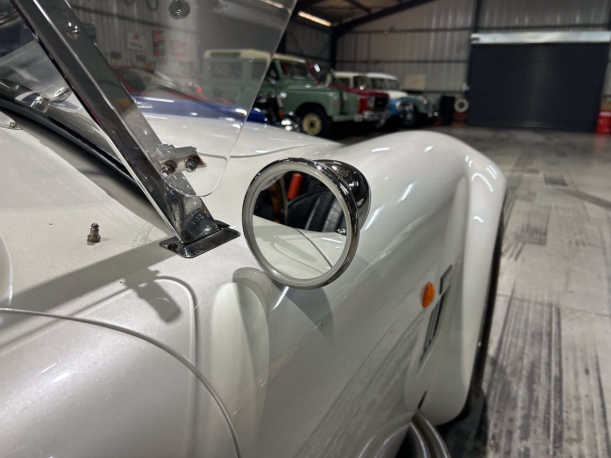Close-up of a vintage white car's chrome side mirror and windshield in a garage, with other classic cars visible in the background.