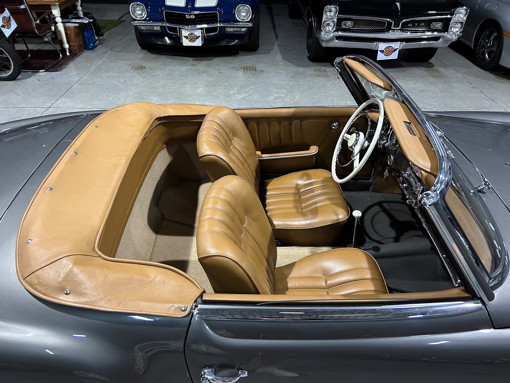Tan leather convertible car interior with cream steering wheel, exposed chrome dashboard, and rear seating in a showroom.
