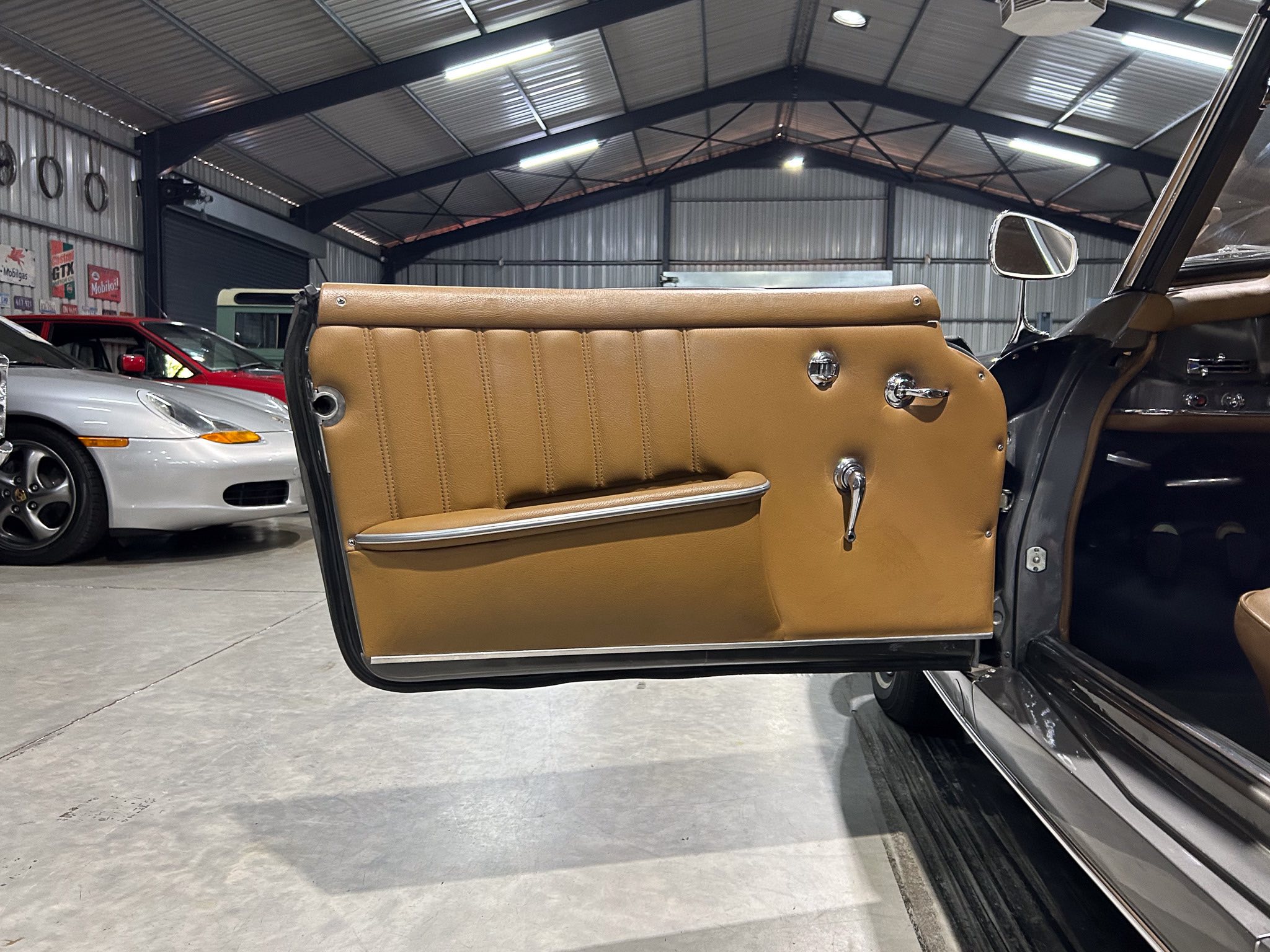 Open car door showing tan leather interior in a garage with other classic cars in the background