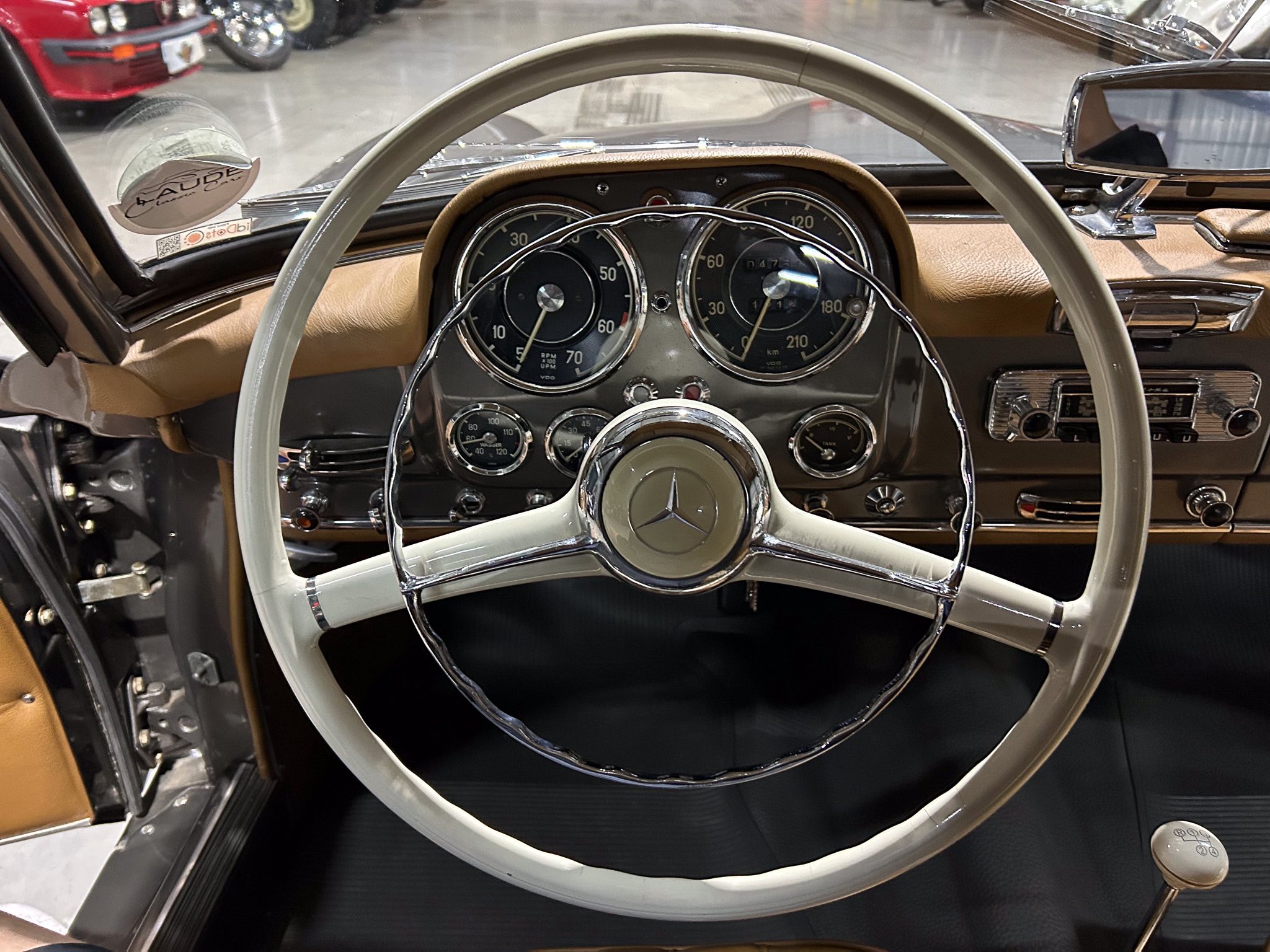 Classic car interior: large white steering wheel with Mercedes emblem, brown leather dash, and cluster of analog gauges.