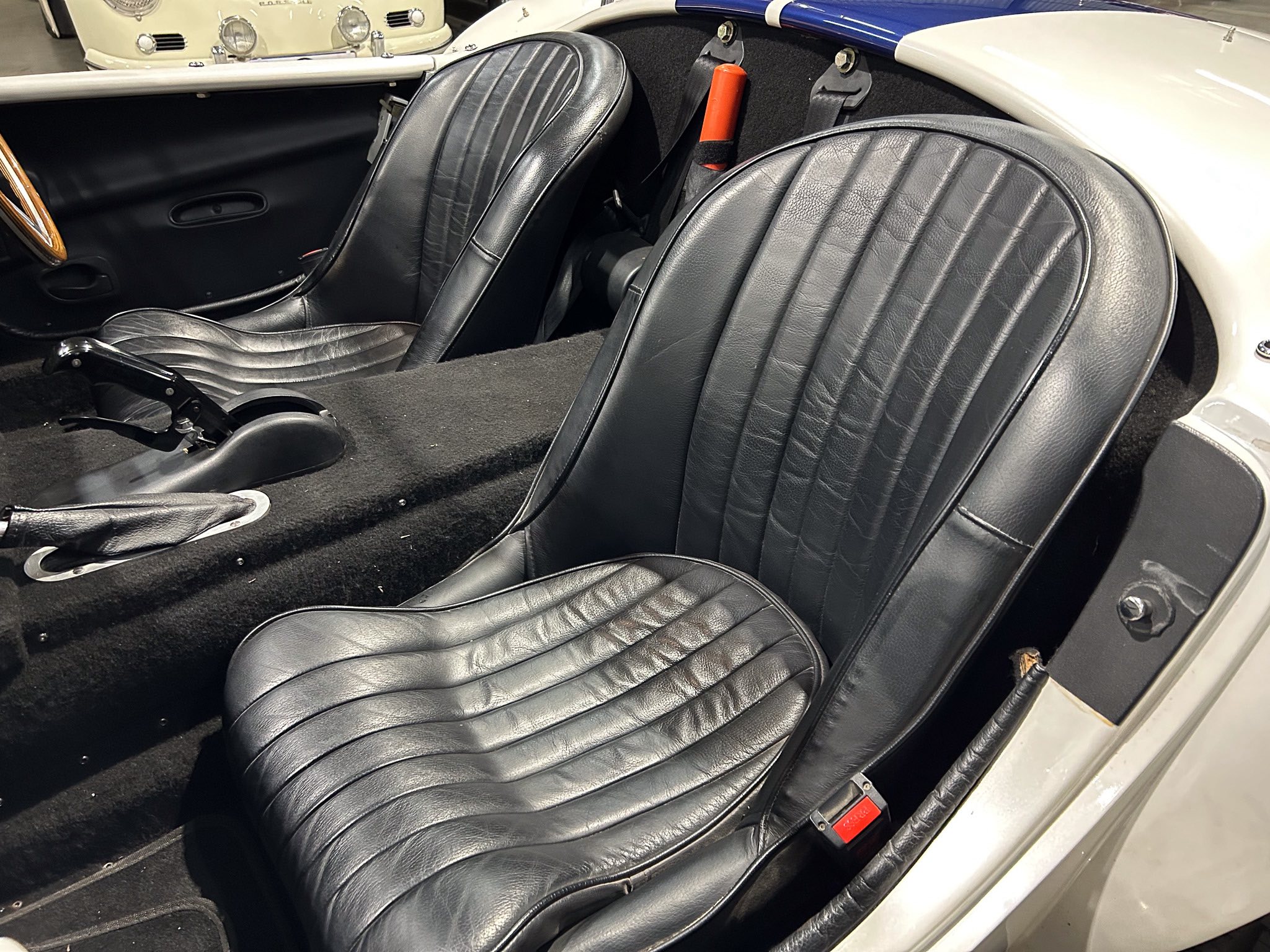 Black leather vintage car interior with two bucket seats and a floor-mounted gear lever in a classic roadster. Light-colored exterior body visible at the door frame; red seatbelt buckle nearby.