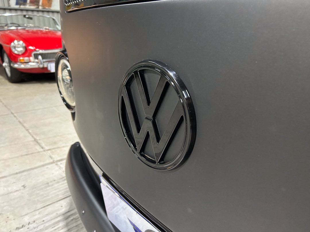Close-up of a black Volkswagen emblem on the front of a dark vehicle, with a red classic car in the background.