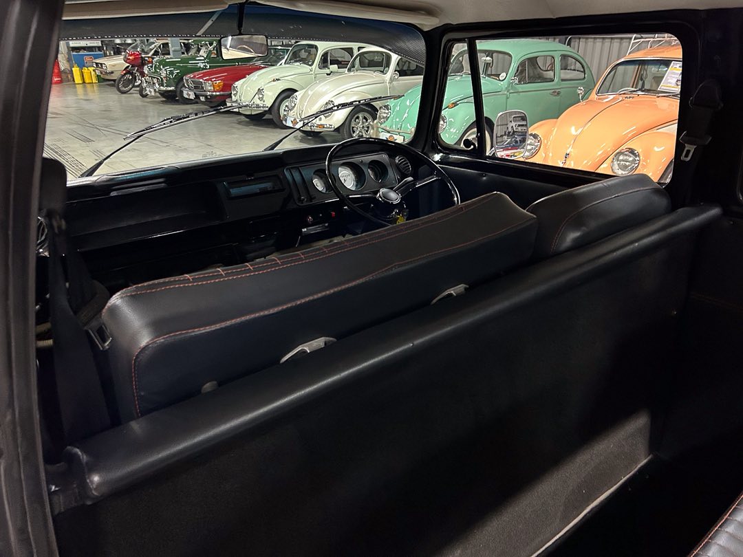 Interior of a black vintage car with leather seats, showing the dashboard and steering wheel, parked in a showroom with classic colorful cars outside the window