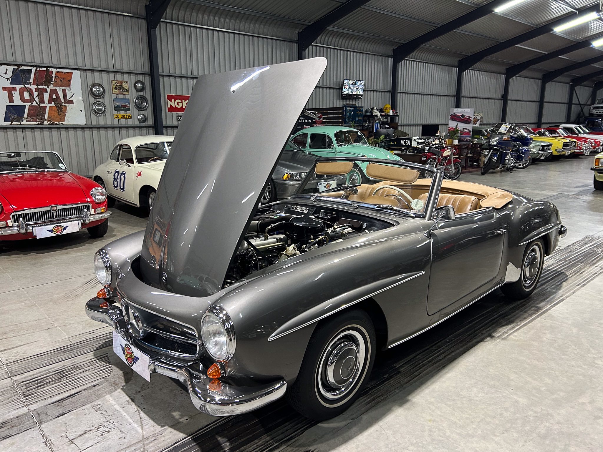 Classic car showroom with a gray convertible in the foreground hood raised, surrounded by rows of vintage cars inside a metal-walled building.