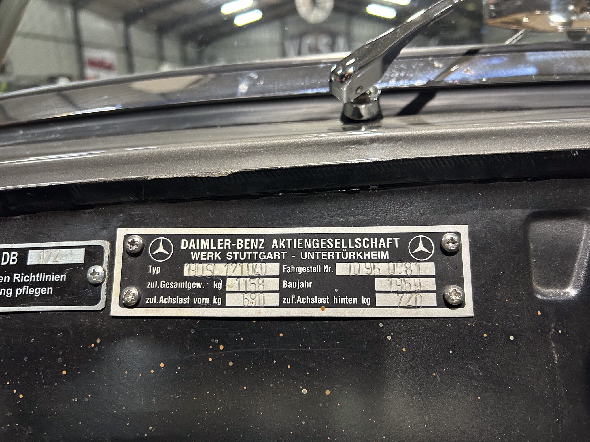 Close-up of a metal vehicle data plate mounted on a car, with screws and embossed text in German identifying the maker (Daimler-Benz) and specifications.