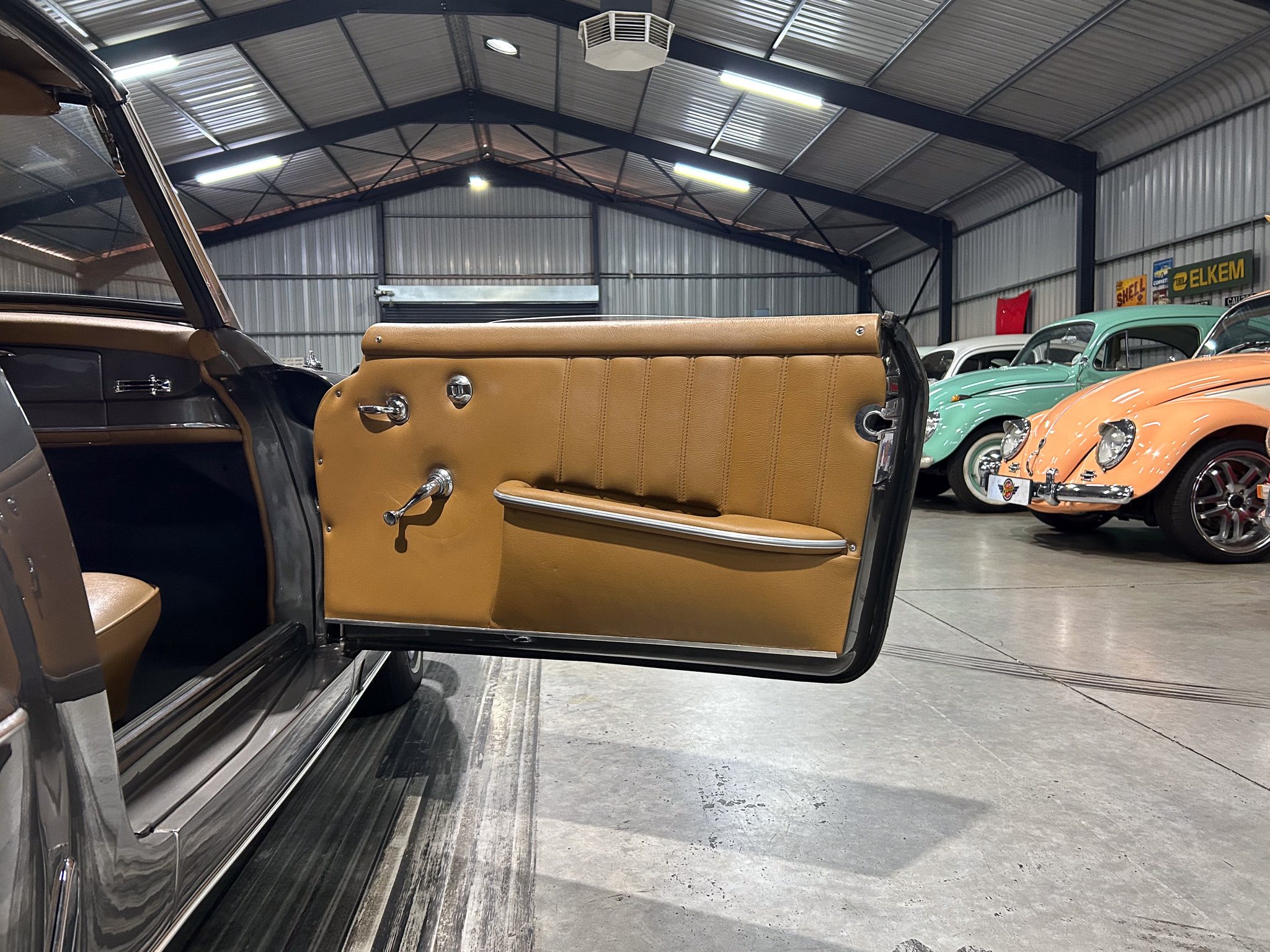 Open door of a tan vintage car showing its leather interior, with pastel-colored classic cars in a metal showroom background.