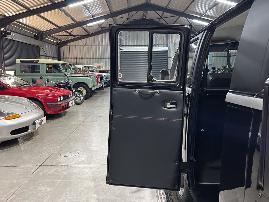 Open car door of a black vehicle in a large indoor showroom filled with vintage cars and a motorcycle in the background, under a metal roof.