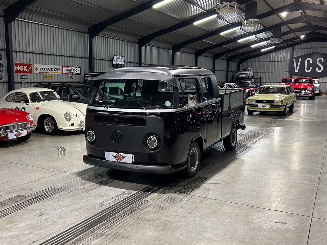 Black vintage Volkswagen Type 2 van in a large indoor showroom surrounded by classic cars on display.