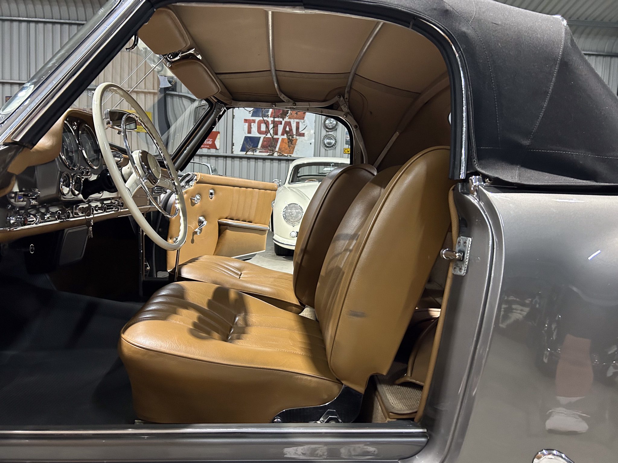 Vintage car interior with tan leather seats and a light-colored steering wheel, seen through an open door with the soft top retracted.