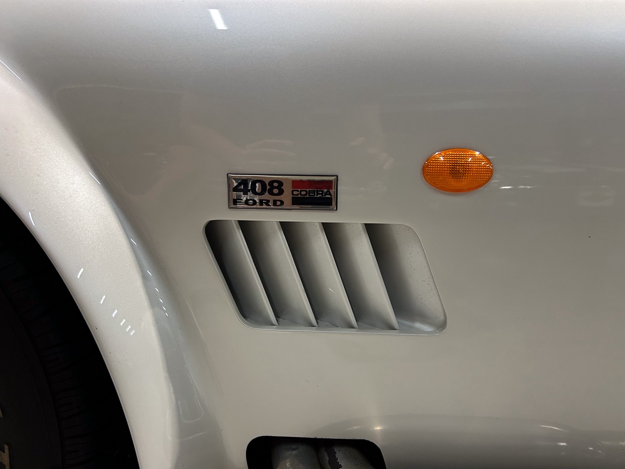 Close-up of a light-gray vehicle front bumper showing an orange side marker light and a chrome badge that reads '408 FORD COBRA' above the grill slats.