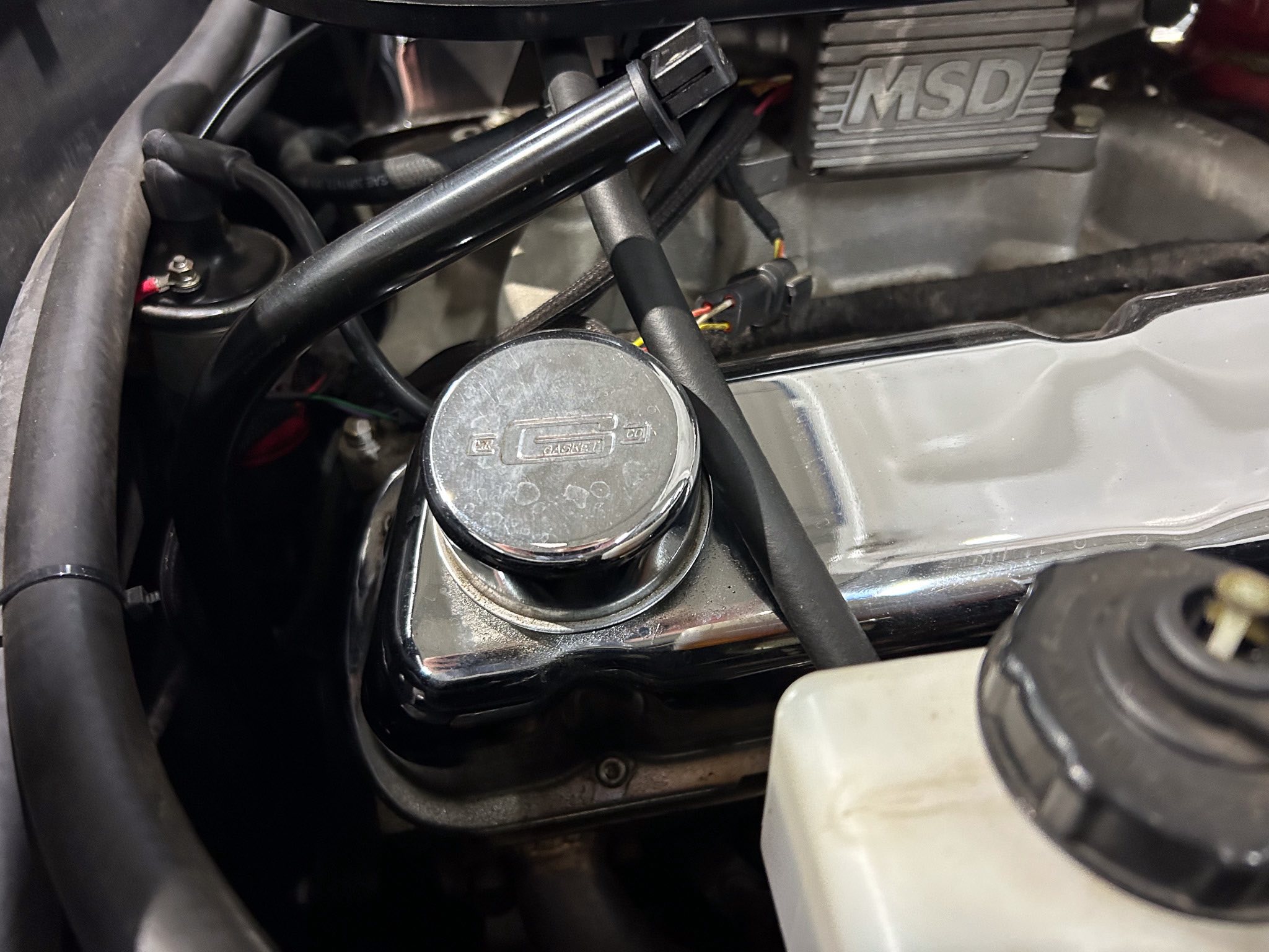 Close-up of a vehicle engine bay with an MSD ignition box, wiring, and hoses visible near a chrome cap.