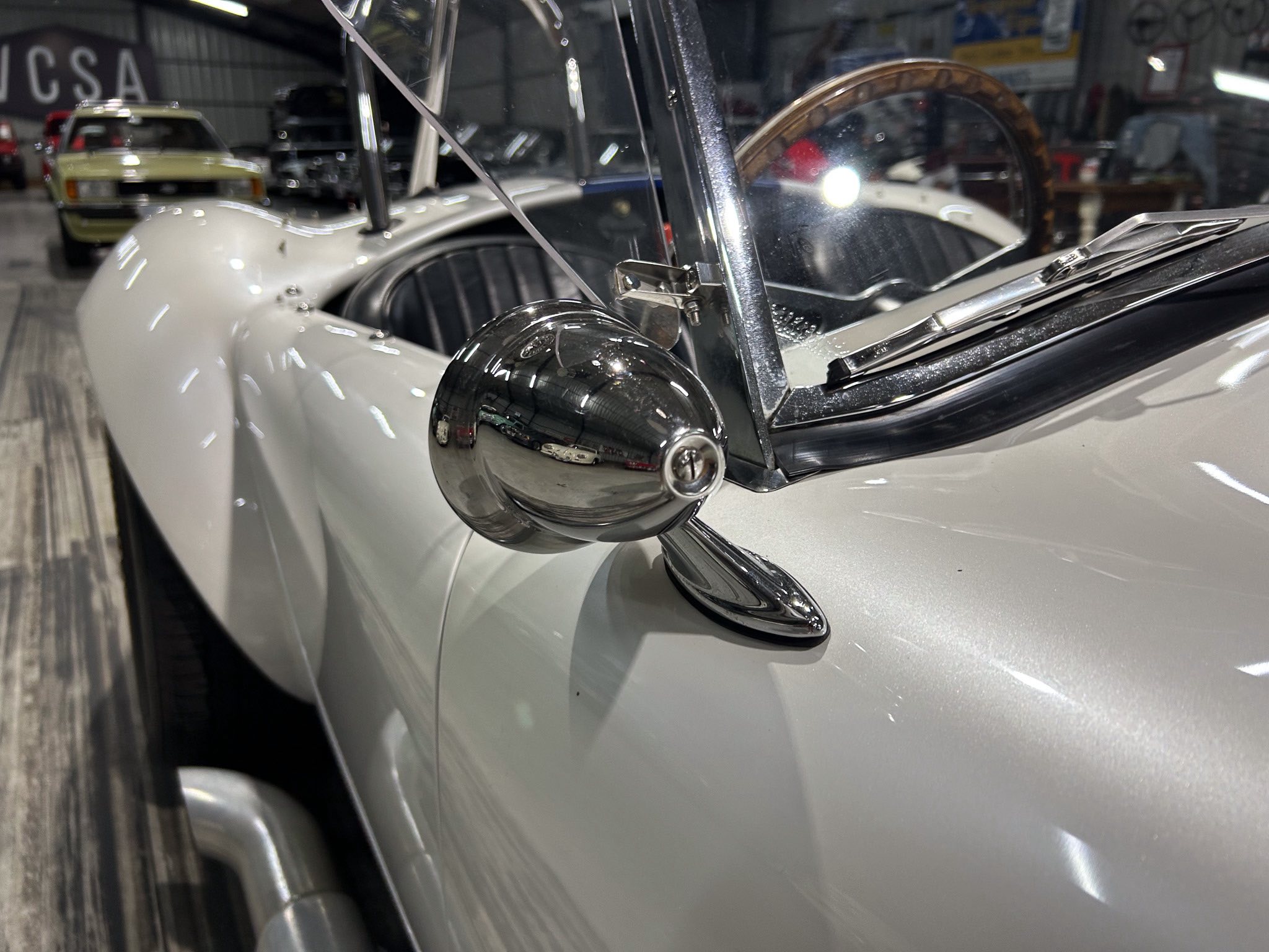 Close-up of a vintage car's chrome side mirror and sharp windshield with a workshop interior in the background.