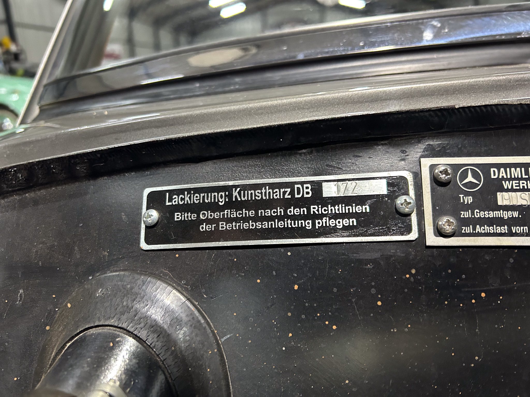 Close-up of a vehicle door jamb with a metal label displaying German maintenance text, including 'Lackierung: Kunstharz DB', and a Mercedes-Benz emblem nearby.