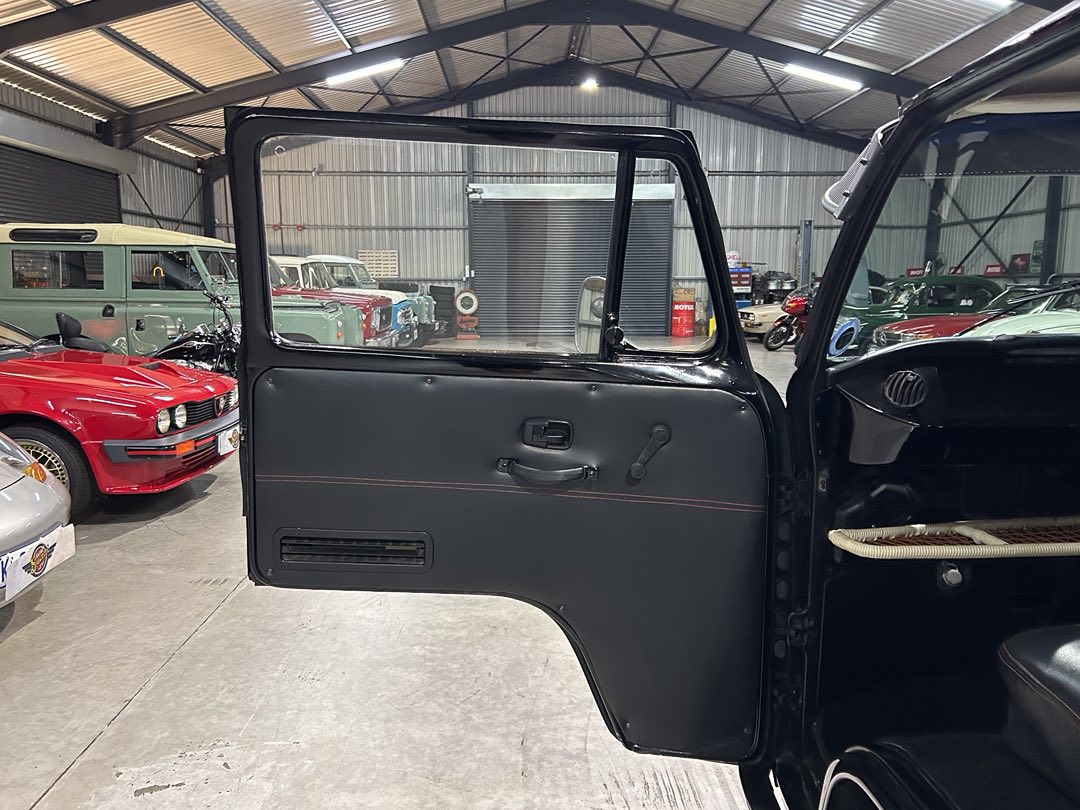 Open black vintage car door showing a gray interior panel with a manual window crank inside a warehouse filled with classic cars