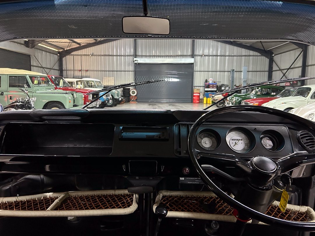 Classic car interior: black dashboard and large steering wheel, with vintage gauges visible, seen from the driver's seat inside a workshop filled with antique vehicles.