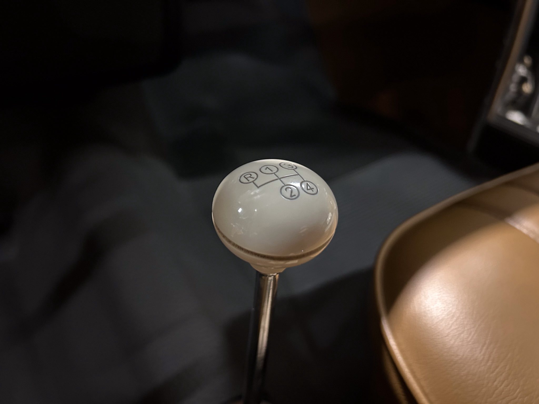 Close-up of a white manual gear shift knob showing gear positions R, 1, 2, 3, 4 with a metal rod in a dark car interior