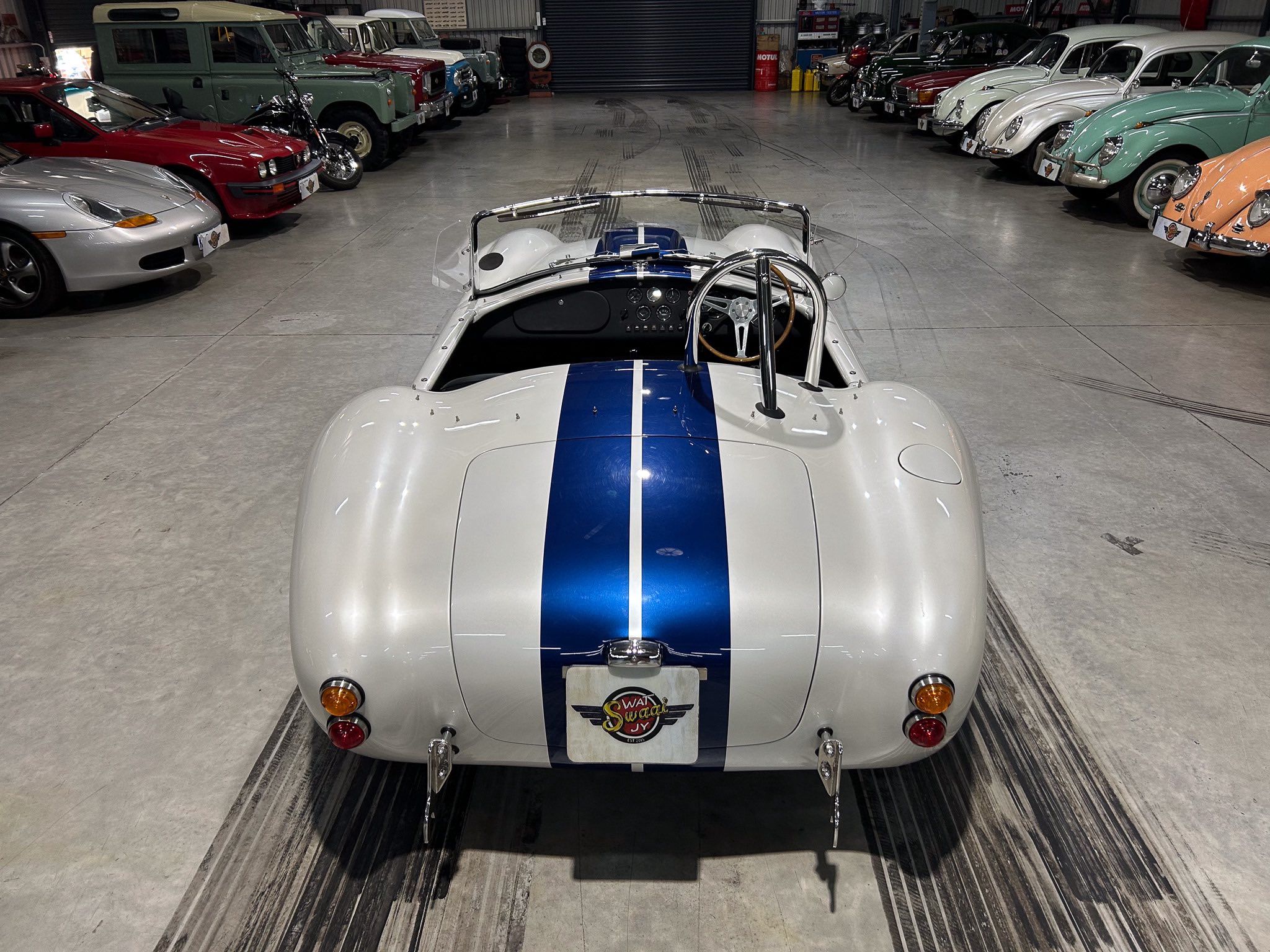 Rear view of a white vintage race car with a blue racing stripe, parked in a showroom flanked by rows of classic cars and vans.