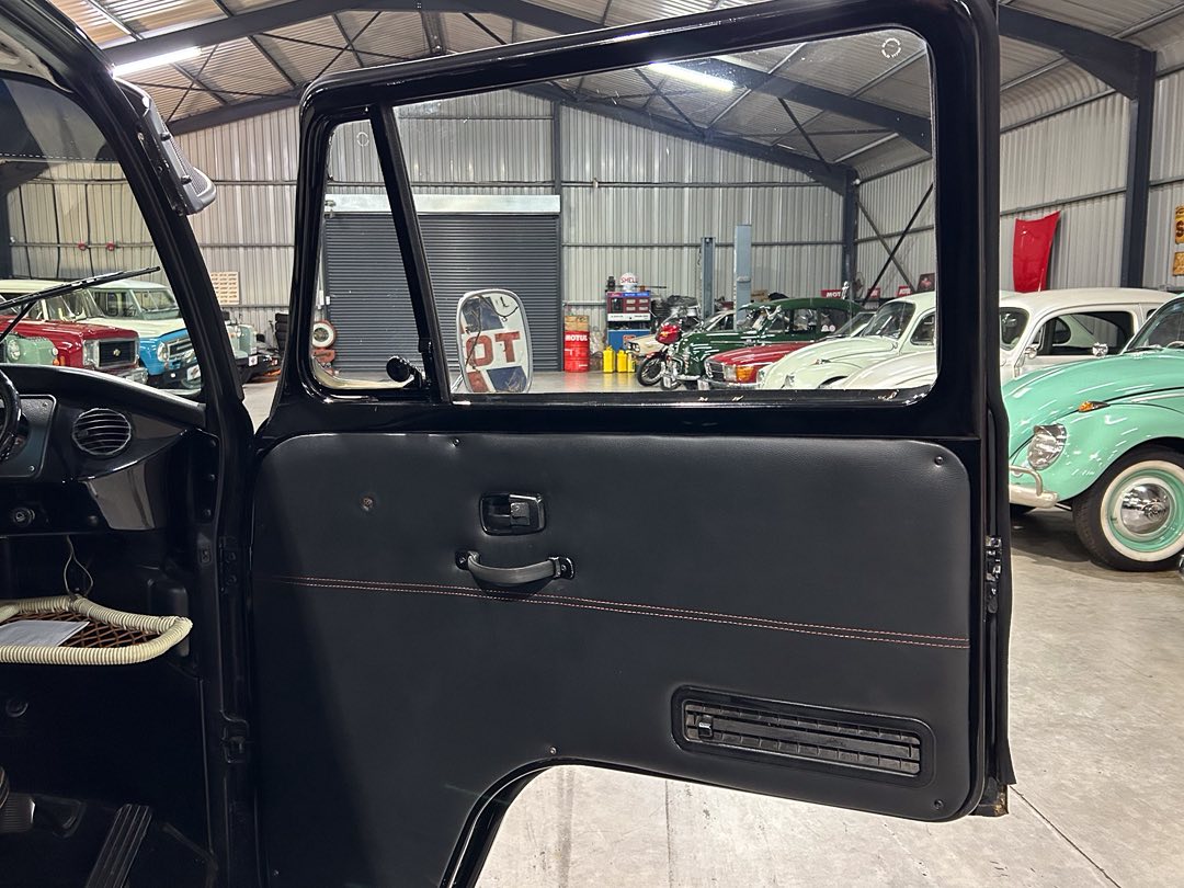 Open vintage car door showing the black interior panel with red stitching, in a showroom with classic cars visible in the background.