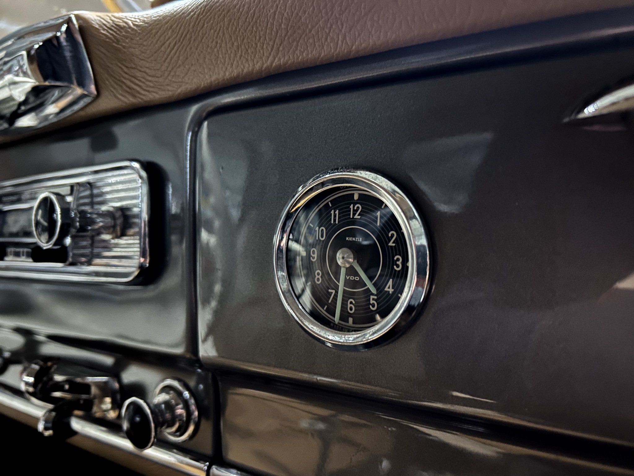 Close-up of a classic car dashboard featuring a round chrome-trimmed analog clock and surrounding control knobs on a dark panel.